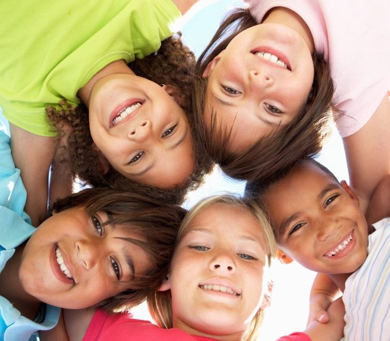 A group of five smiling young people in a huddle looking down at a camera on the floor.